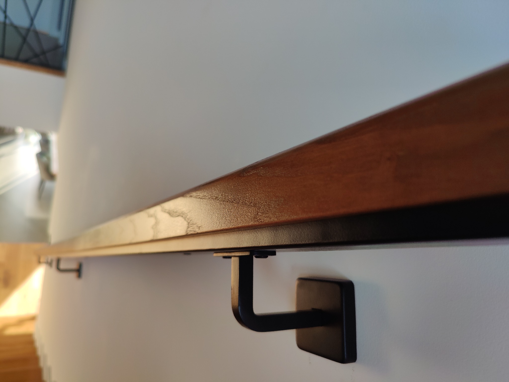 Drewniana poręcz przymocowana do ściany za pomocą czarnych, metalowych wsporników. Widoczny fragment schodów w tle.