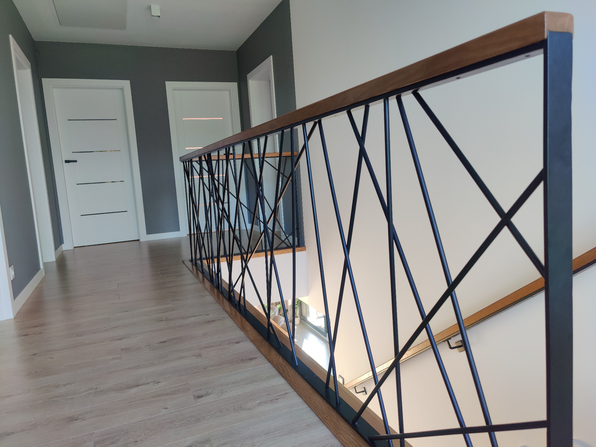 Nowoczesna balustrada wewnętrzna z czarnych, krzyżujących się prętów i drewnianym wykończeniem, widoczna z perspektywy podłogi, z białymi drzwiami w tle.