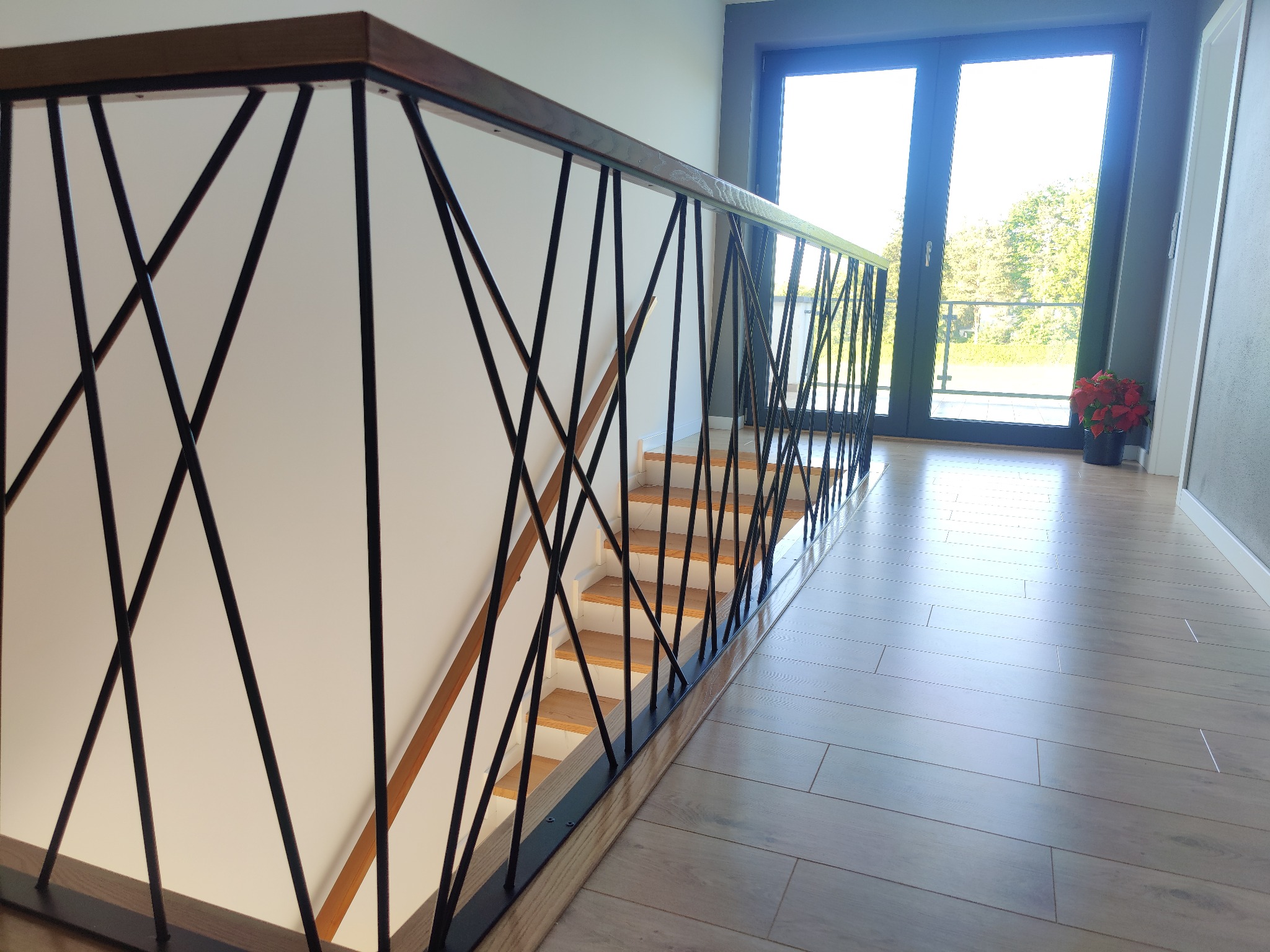 Nowoczesna balustrada schodowa z czarnych, ukośnych prętów i drewnianą poręczą, widok z góry na schody z drewnianymi stopniami i jasną podłogą w tle drzwi balkonowe.