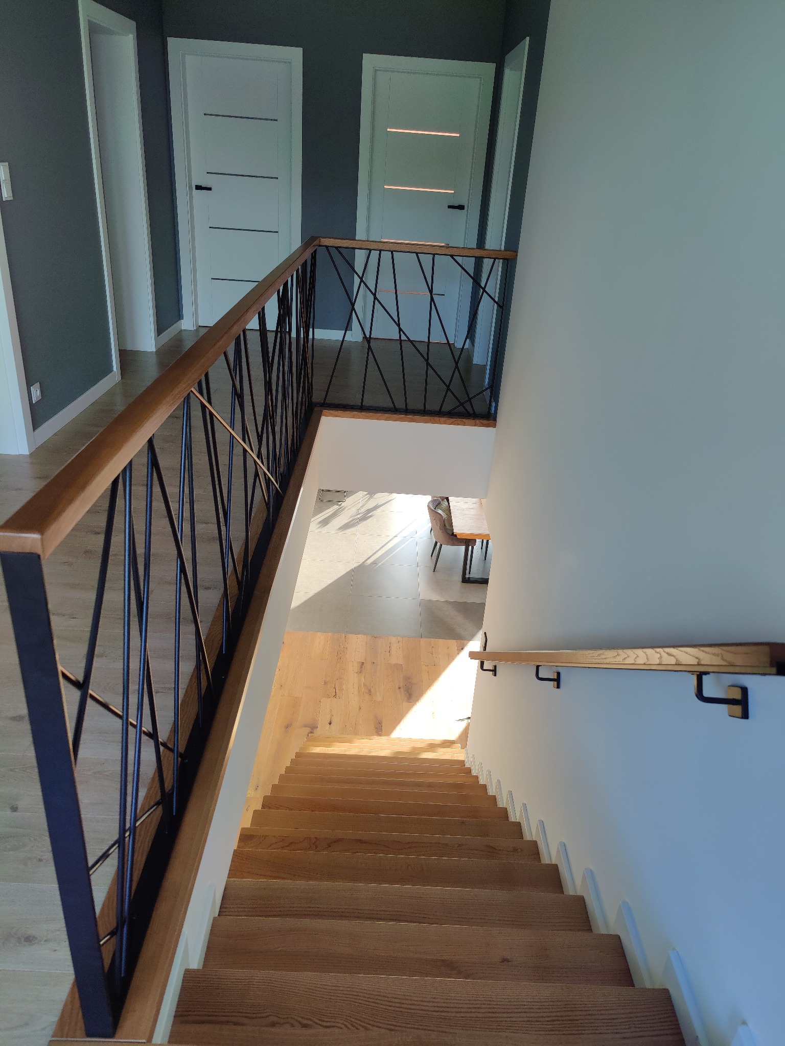 Drewniane schody z czarną, geometryczną balustradą i drewnianą poręczą, widok z góry, w tle białe drzwi i jasne podłogi.