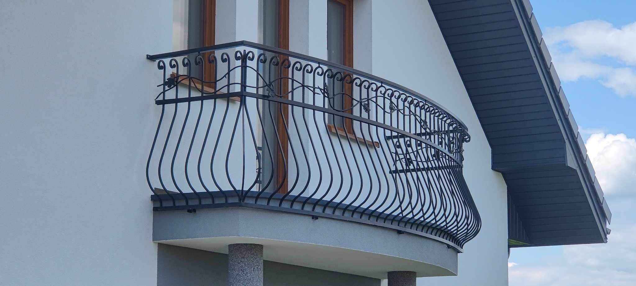 Elegancka, czarna, metalowa balustrada balkonowa z dekoracyjnymi motywami roślinnymi, zamontowana na balkonie budynku z białymi ścianami i brązowymi ramami okiennymi, widok z dołu.