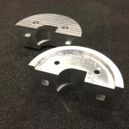 Dwa precyzyjnie obrobione, półokrągłe elementy metalowe z widocznymi śladami frezowania i otworami, leżące na ciemnym, teksturowanym podłożu.