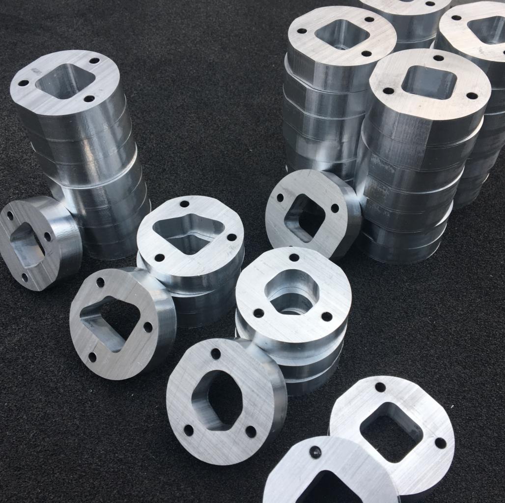 Stosy srebrnych, metalowych elementów z otworami o różnych kształtach (kwadrat, romb, sześciokąt) i otworami na śruby, ułożone na ciemnoszarym, teksturowanym tle.