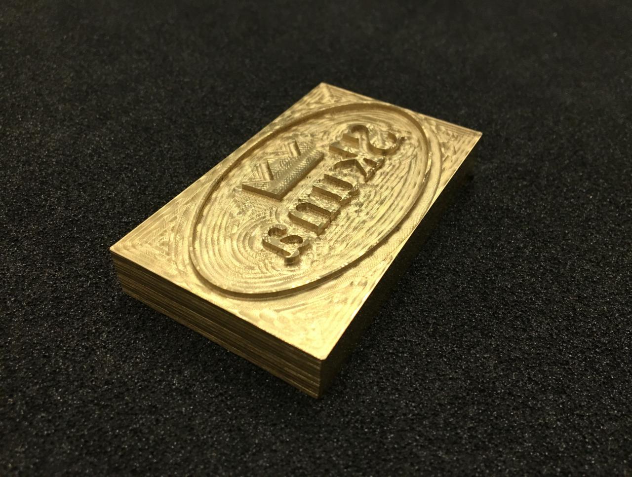 Mosiężna matryca z wygrawerowanym logo wykonana w Poznaniu, leżąca na czarnym, teksturowanym tle. Widoczne ślady obróbki.