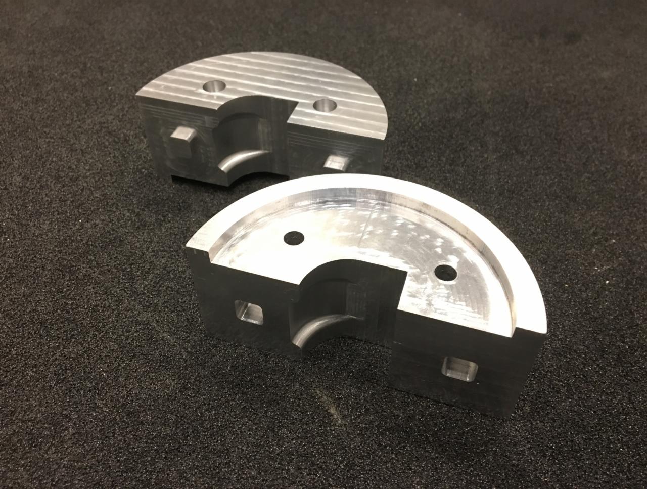 Dwa precyzyjnie obrobione, półokrągłe elementy metalowe z widocznymi śladami frezowania i otworami, leżące na ciemnym, teksturowanym podłożu.