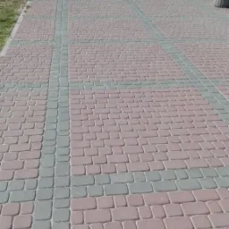 Szeroki chodnik z czerwonej kostki brukowej z szarymi pasami, prowadzący do budynku z ławkami i koszami na śmieci.