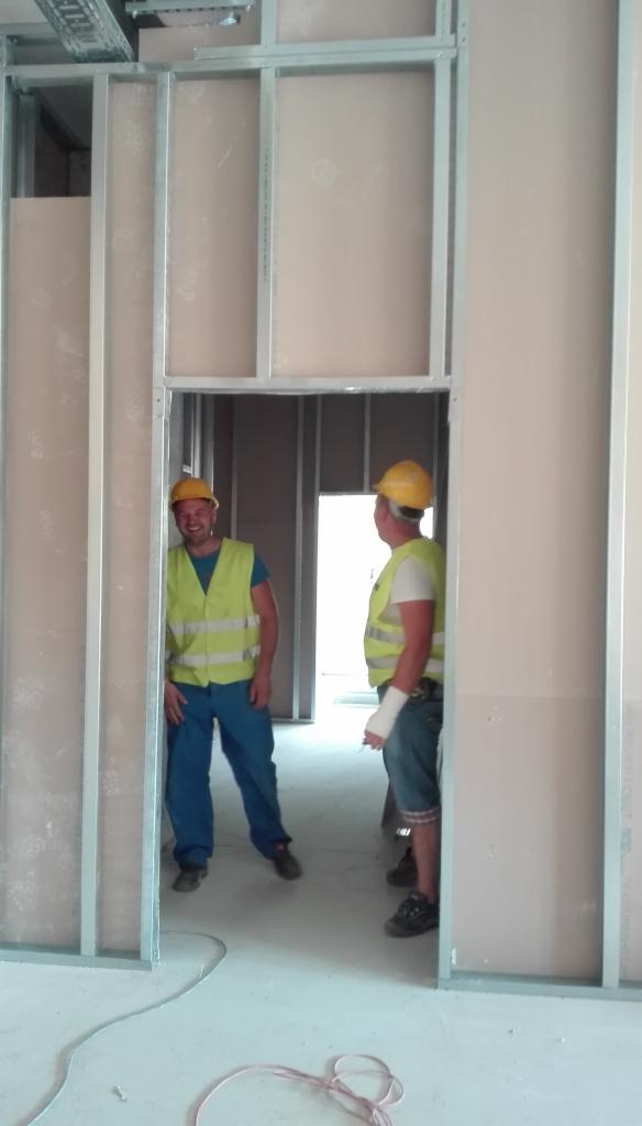 Montaż ścian działowych z płyt gipsowo-kartonowych. Dwóch pracowników w kaskach i kamizelkach odblaskowych w trakcie pracy. Widoczna metalowa konstrukcja szkieletowa ścian.