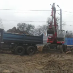 Ciężarówka z wywrotką wypełniona ziemią i czerwona koparka kołowa w trakcie załadunku ziemi na placu budowy, widoczne ślady opon w błocie.