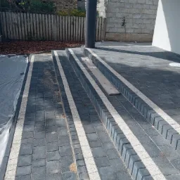 Schody i ścieżka z ciemnoszarej kostki brukowej z białymi akcentami, prowadzące do budynku z widocznym fragmentem elewacji i filarem, w tle drewniany płot i budynek z czerwonym dachem.