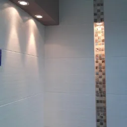 Wykończona ściana łazienki z beżowymi płytkami imitującymi drewno, pionowy pas mozaiki i oświetlenie punktowe w suficie podwieszanym i w pasie mozaiki.