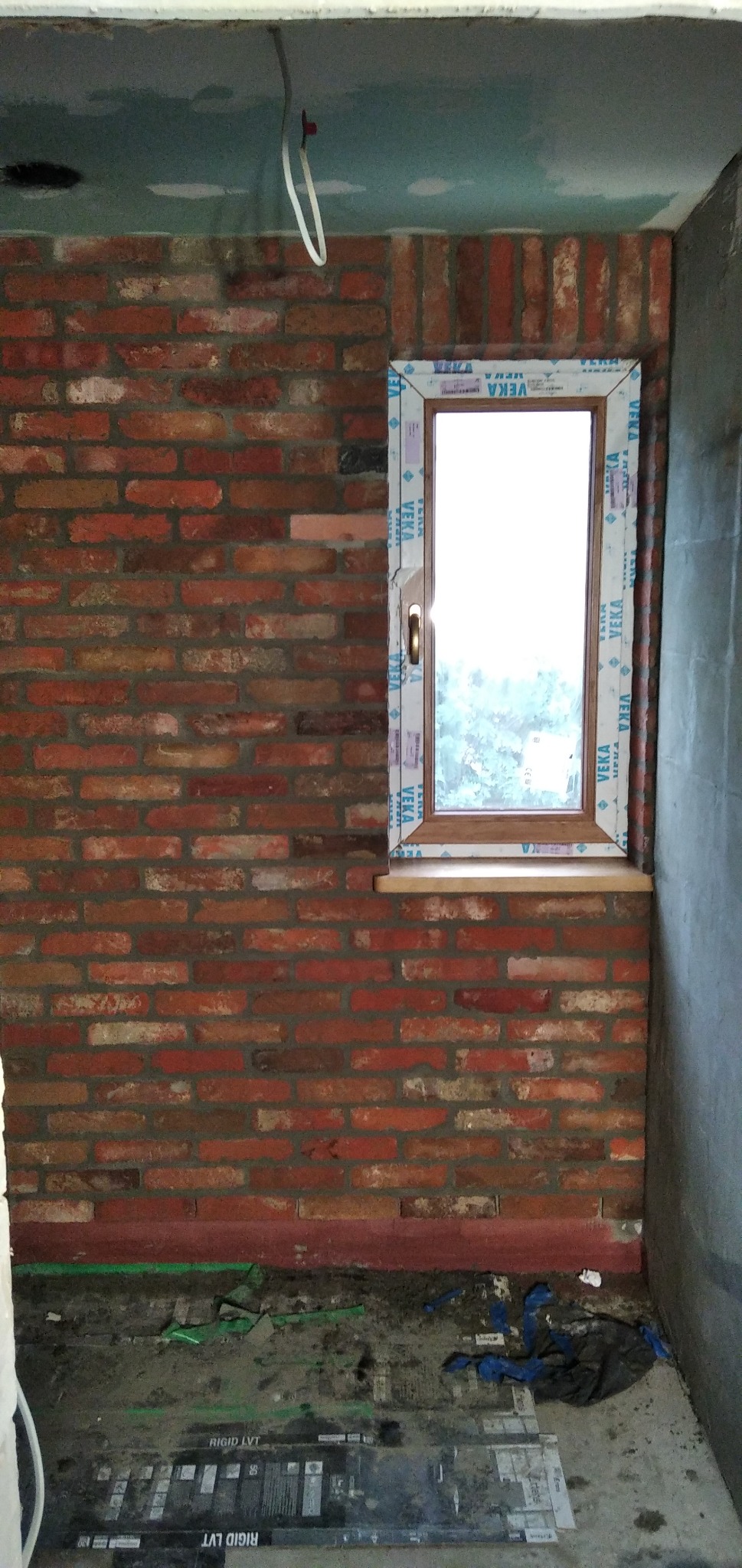 Ceglana ściana z nowym oknem PCV marki VEKA, widoczne ślady remontu na podłodze i surowy sufit z wystającym przewodem.