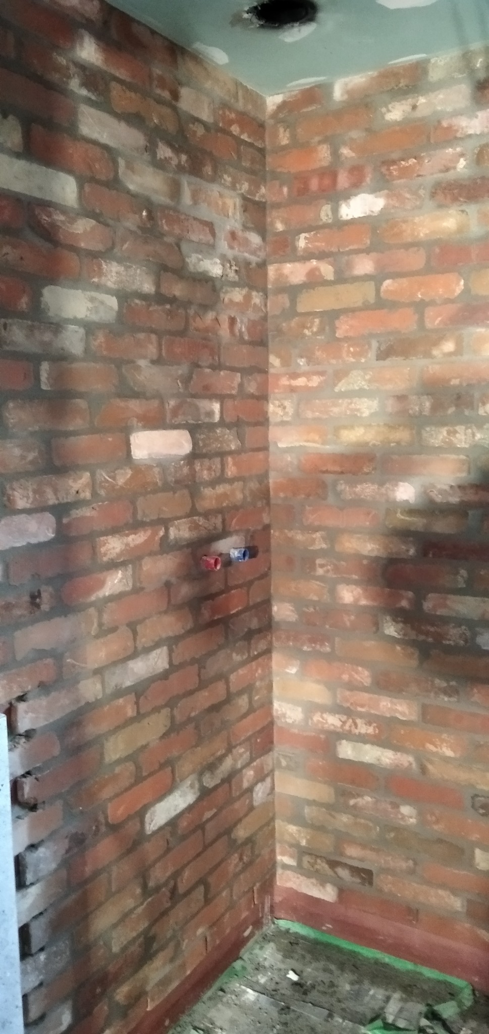 Fragment pomieszczenia w trakcie wykańczania z widoczną ścianą z cegły klinkierowej, wystającymi przyłączami wodnymi i niedokończonym sufitem podwieszanym.