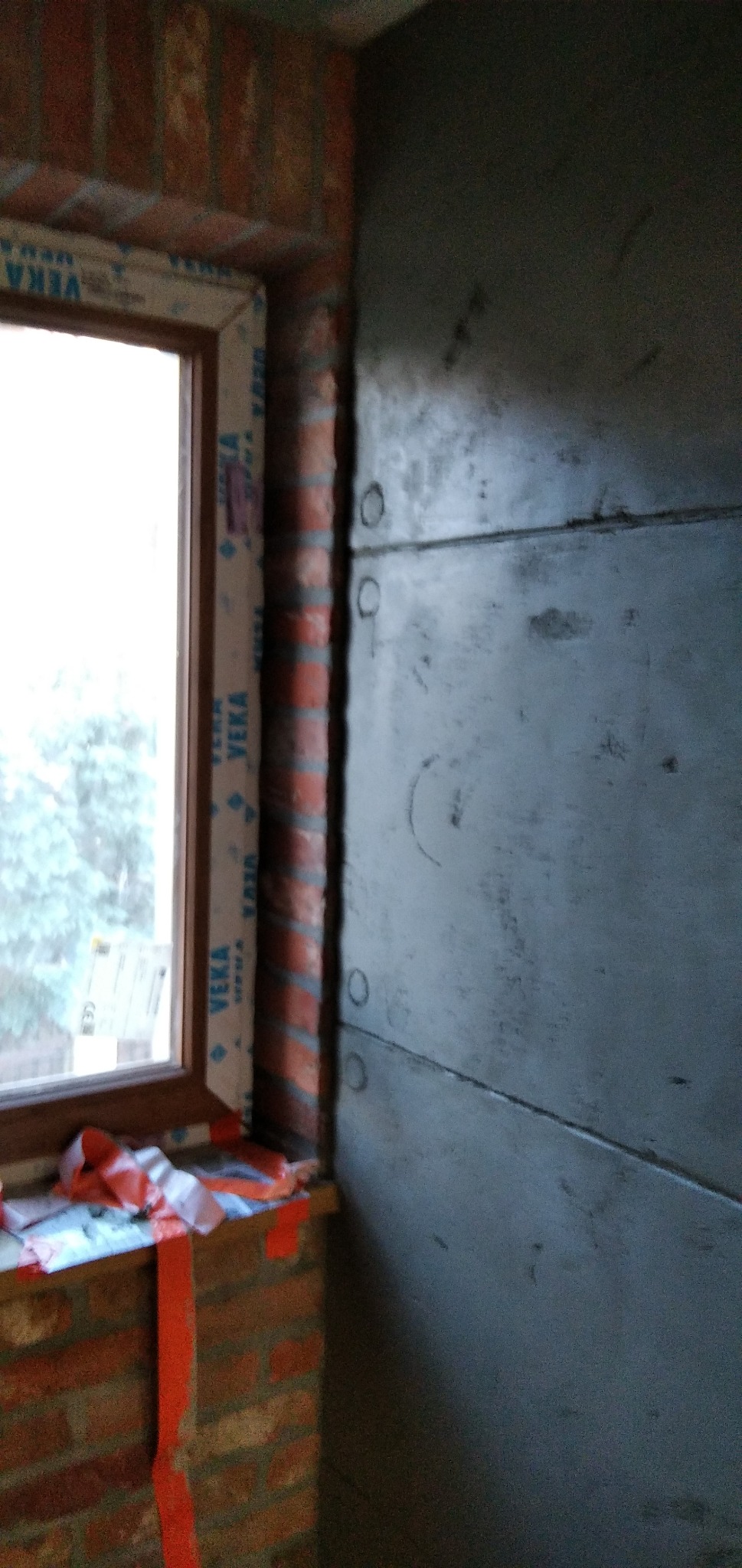 Surowa ściana z płyt gipsowo-kartonowych mocowanych na stelażu, widoczny fragment okna z ceglanym obramowaniem i taśmą zabezpieczającą na parapecie.