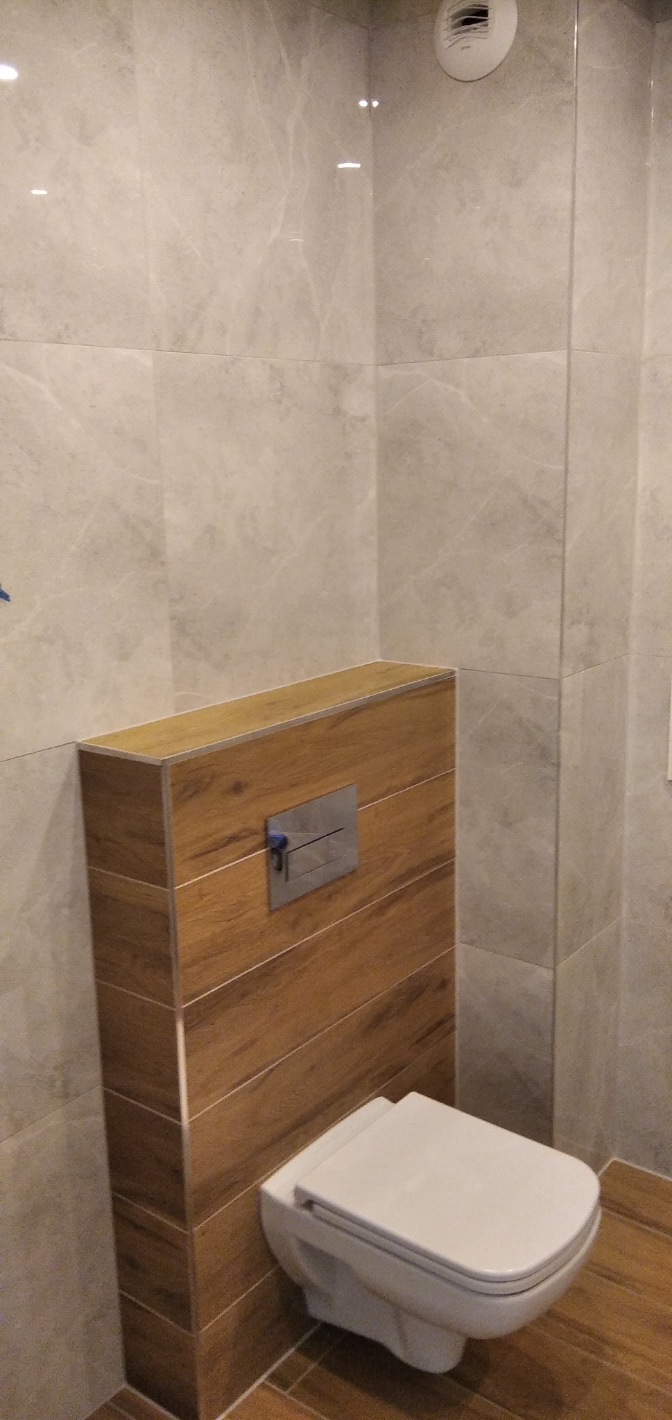 Wisząca toaleta z zabudową z płytek imitujących drewno, na tle ściany wyłożonej szarymi płytkami marmurkowymi, widoczny przycisk spłukiwania.