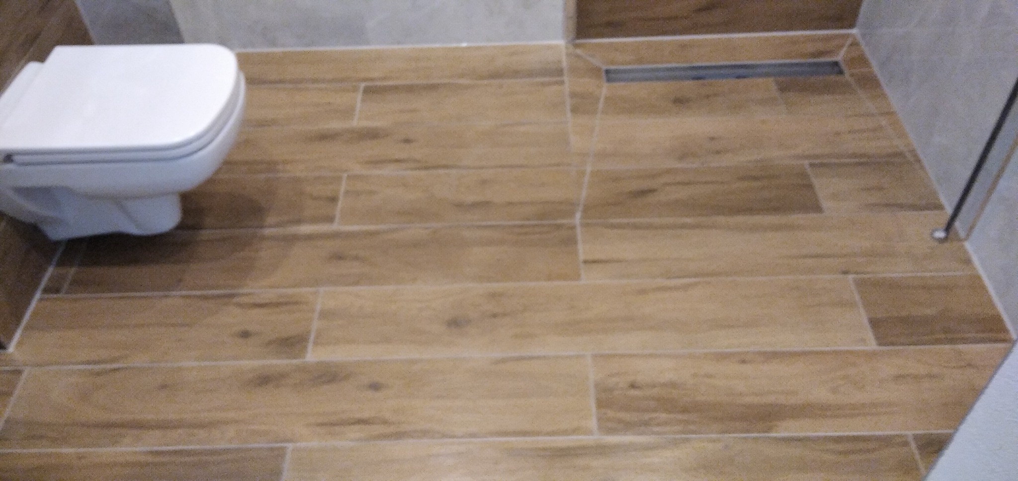 Wykończona łazienka z drewnopodobną podłogą z płytek, wisząca toaleta i odpływ liniowy przy kabinie prysznicowej.