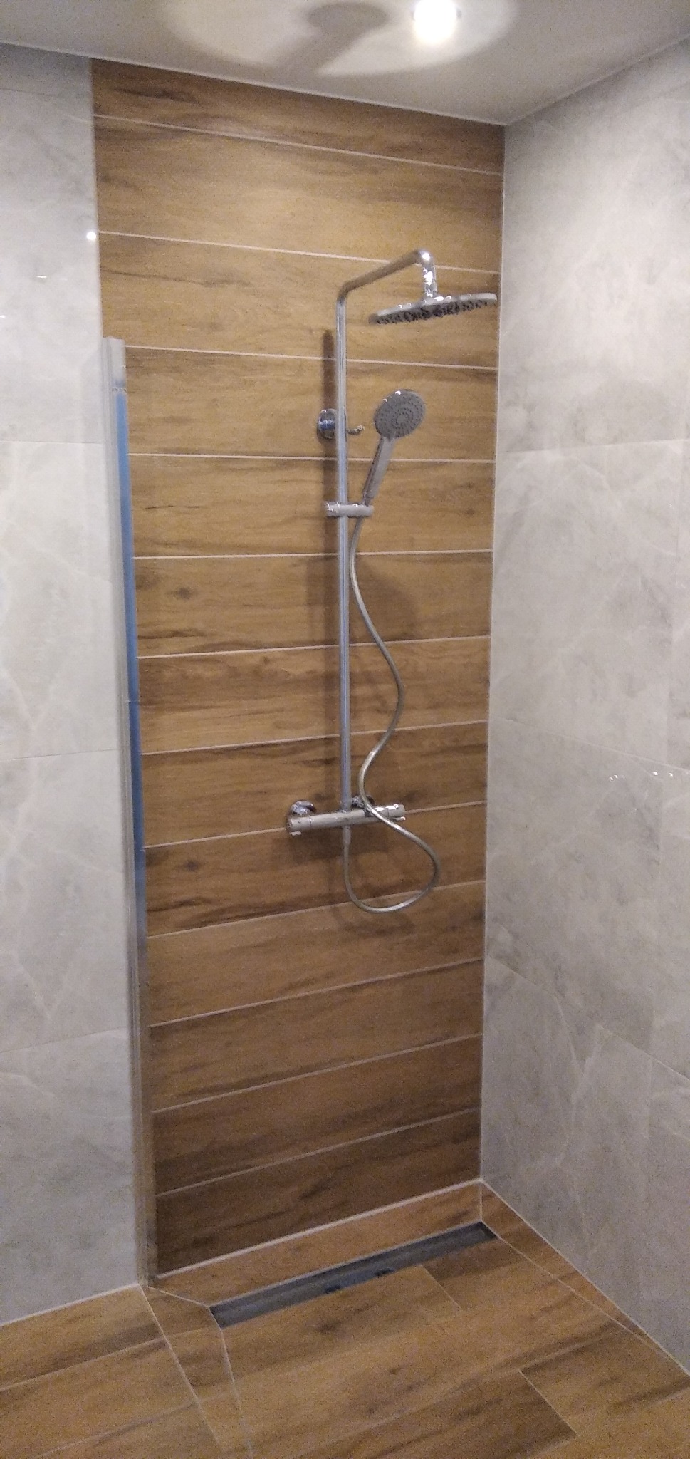 Nowoczesna kabina prysznicowa z deszczownicą i panelami ściennymi imitującymi drewno, wykończona jasnymi płytkami imitującymi marmur, z odpływem liniowym w podłodze.