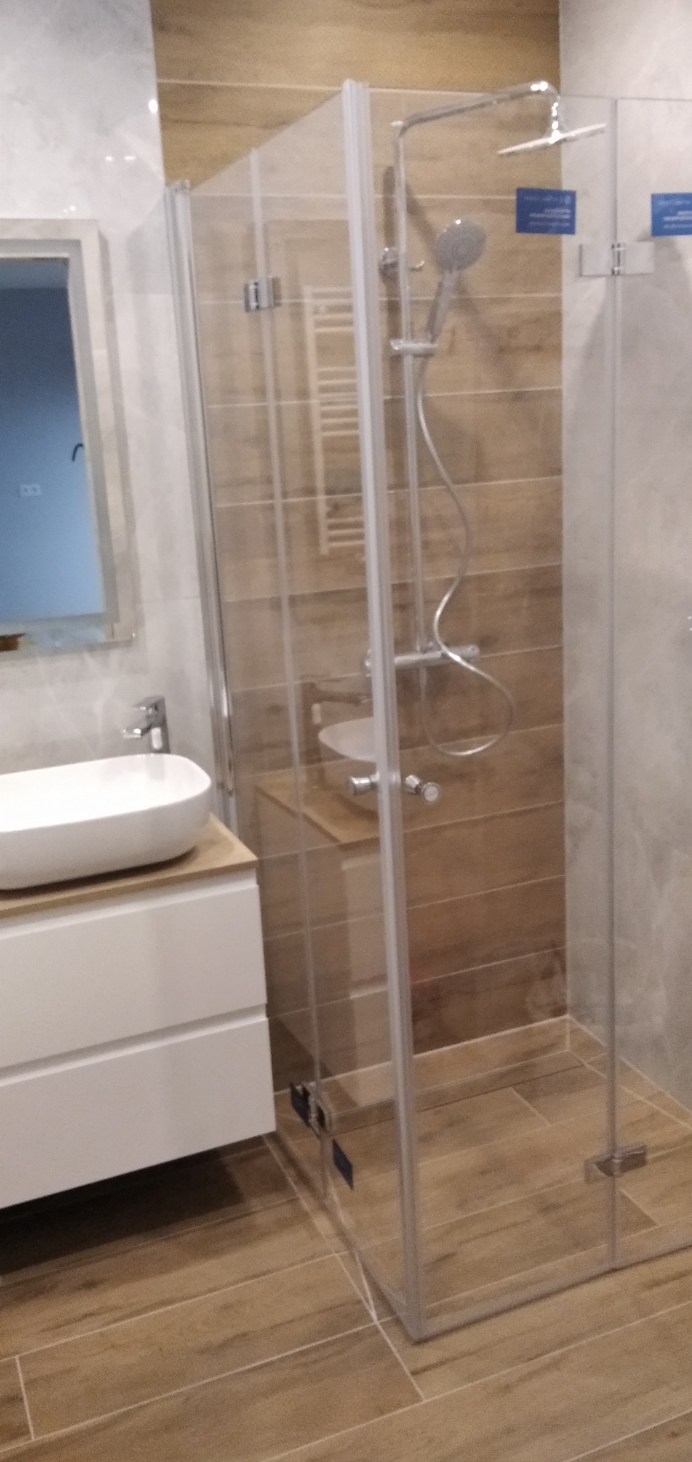 Nowoczesna łazienka z kabiną prysznicową ze szklanymi drzwiami, umywalką nablatową na szafce z białymi frontami i lustrem. Płytki ścienne imitujące drewno. Widoczny także grzejnik drabinkowy...