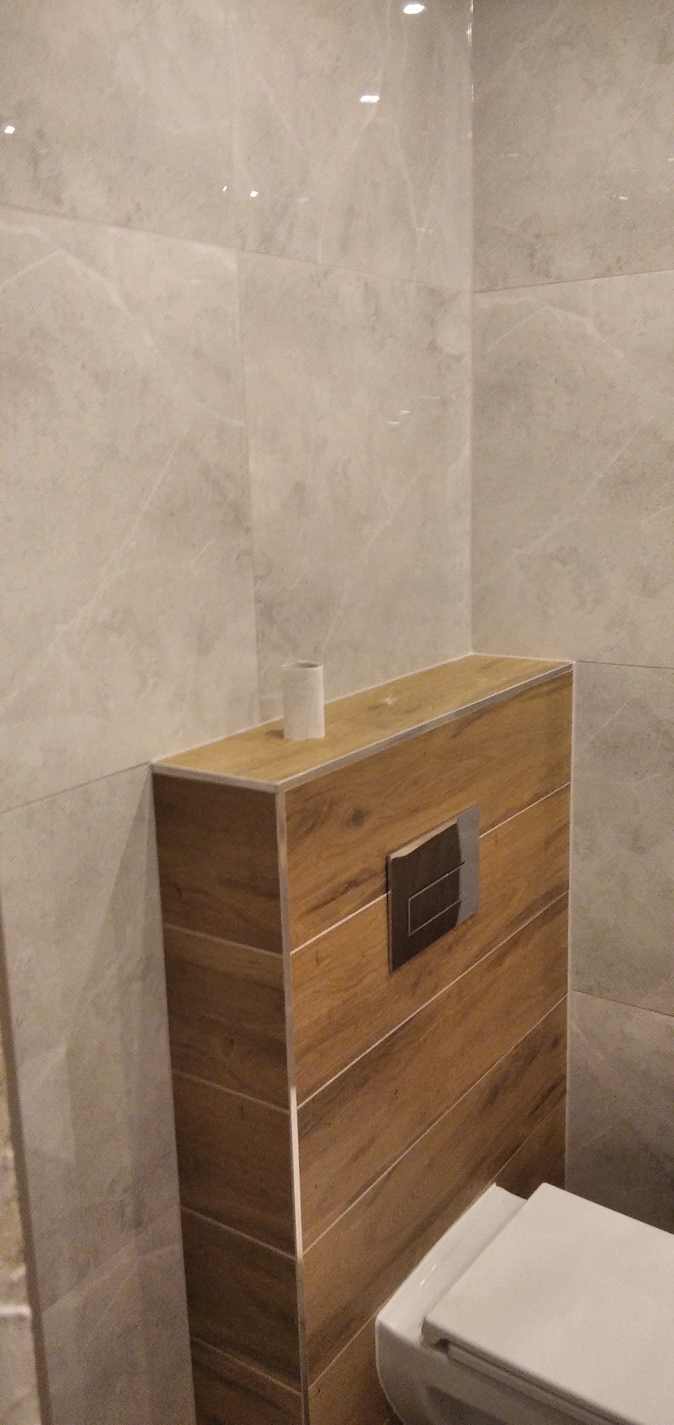 Obudowa stelaża podtynkowego WC wykończona drewnopodobnymi panelami z metalowymi listwami, dopasowana do marmurkowych płytek ściennych, z pustą rolką po papierze toaletowym na górze.