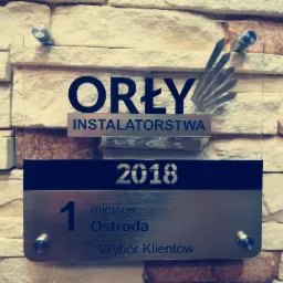 Tablica z nagrodą 'Orły Instalatorstwa' za 1 miejsce w Ostródzie w 2018 roku, przytwierdzona do ściany z kamienia dekoracyjnego.