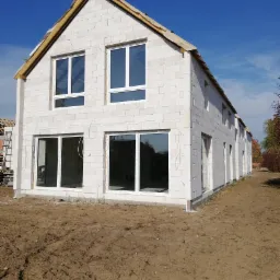 Wznoszony dwukondygnacyjny dom z białych bloczków, widoczne wstawione okna, niezakończone prace dekarskie, teren wokół domu przygotowany pod dalsze prace.
