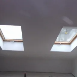 Dwa okna dachowe na poddaszu, widoczne z dołu, po szpachlowaniu i malowaniu. Widoczne gładkie powierzchnie sufitu i ścian w jasnym odcieniu.