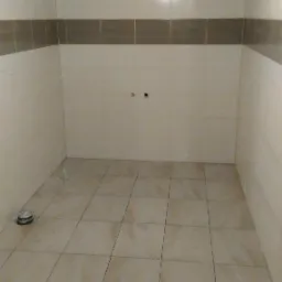 Wykończona łazienka z beżowymi płytkami na podłodze i ścianach, z szarym pasem płytek na ścianach. Widoczny odpływ podłogowy i wisząca żarówka.