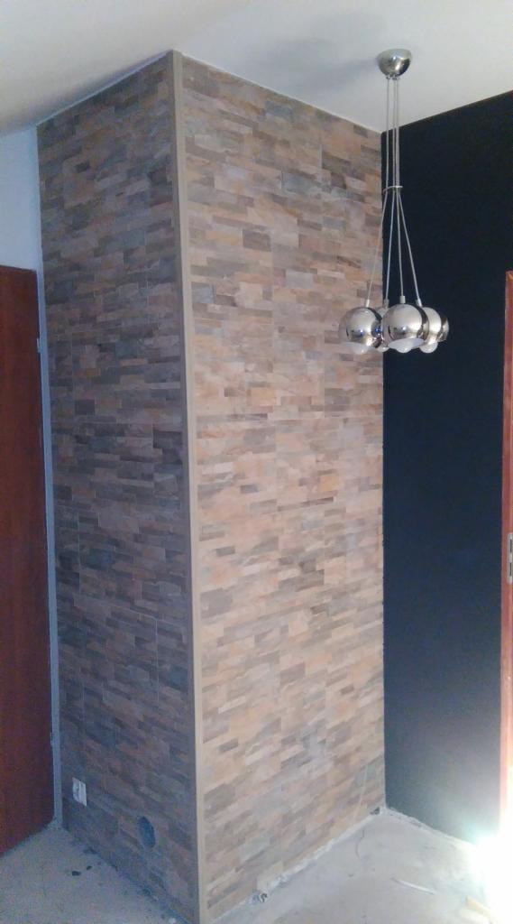 Dekoracyjna ściana z kamiennej okładziny w odcieniach brązu i beżu, oświetlona nowoczesną lampą wiszącą. Widoczny fragment drzwi i ciemna ściana w tle.