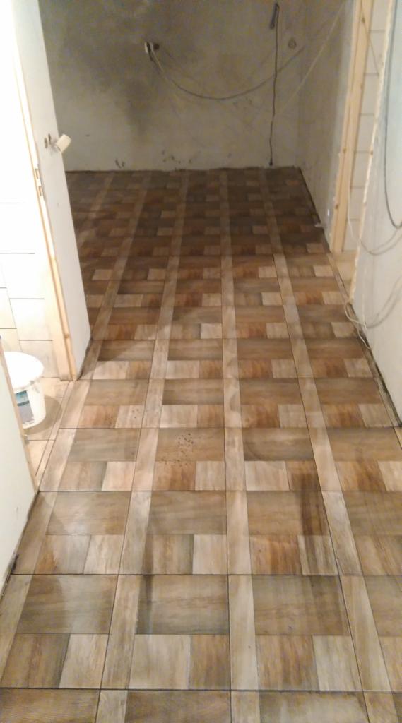 Ułożona podłoga z płytek imitujących drewno w trakcie remontu. Widoczny fragment ściany z instalacją elektryczną i białe drzwi po lewej stronie.