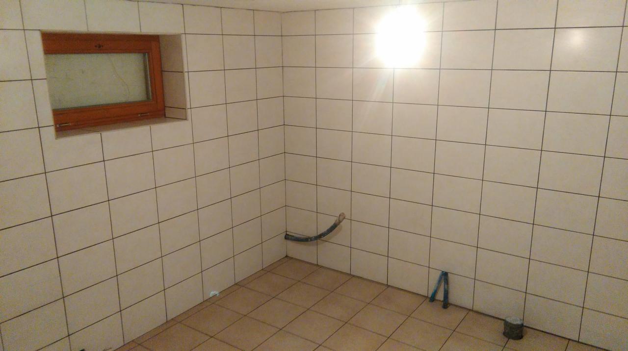 Wnętrze łazienki w trakcie wykończenia: jasne płytki na ścianach i podłodze, małe okno z drewnianą ramą, fragment instalacji wodnej i narzędzia na podłodze.