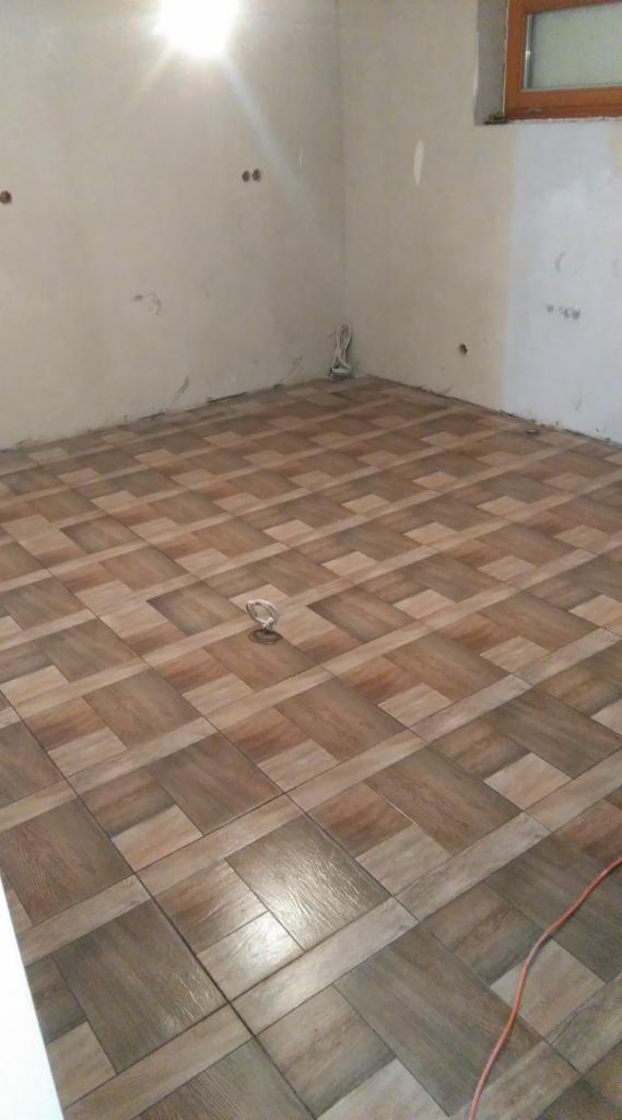 Ukończona podłoga z brązowych, drewnopodobnych płytek ułożonych we wzór. Ściany surowe, w tle okno. Widoczny przewód elektryczny.