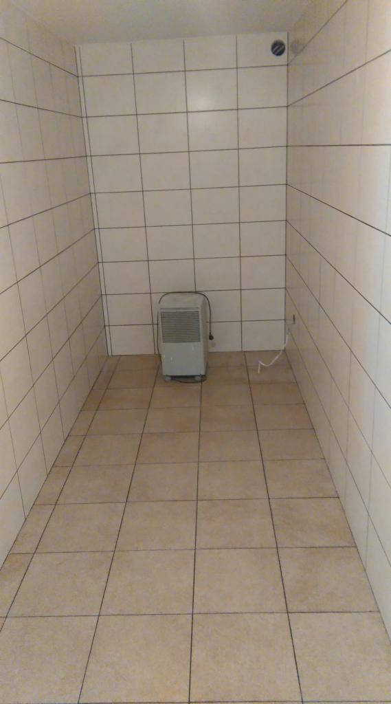 Wykończona łazienka z beżowymi płytkami na podłodze i ścianach. W rogu stoi grzejnik. Widoczny otwór wentylacyjny na suficie i wystający kabel.