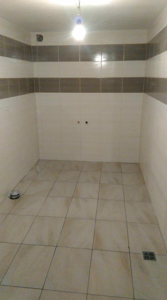 Wykończona łazienka z beżowymi płytkami na podłodze i ścianach, z szarym pasem płytek na ścianach. Widoczny odpływ podłogowy i wisząca żarówka.