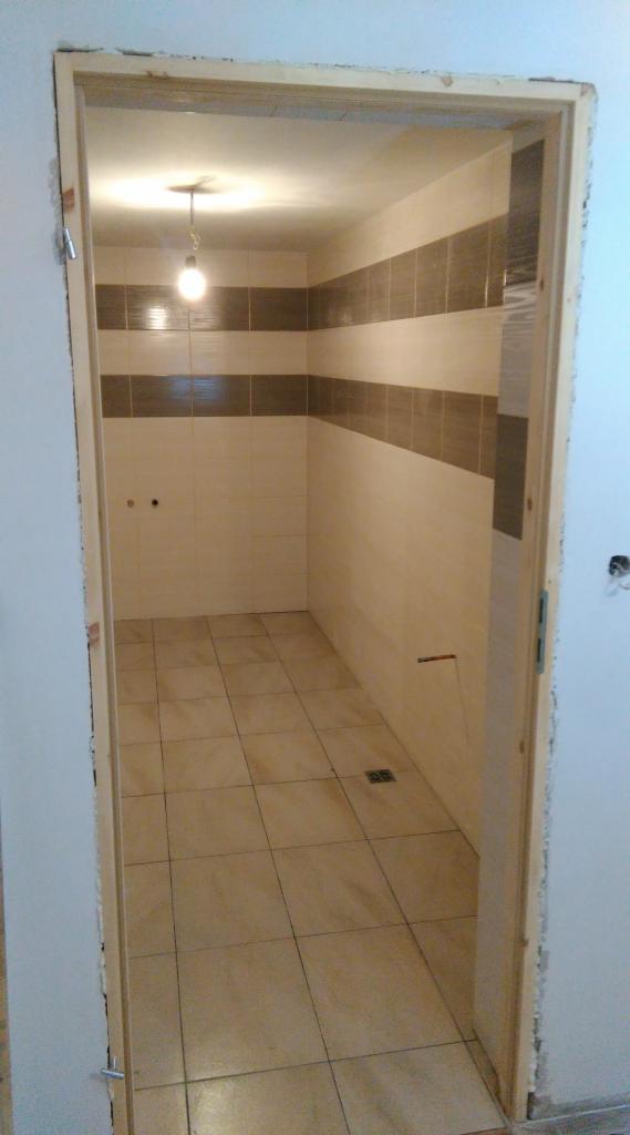 Wykończona łazienka z beżowymi płytkami podłogowymi i pasem brązowych płytek na ścianie, widoczna zza surowej ościeżnicy drzwiowej.