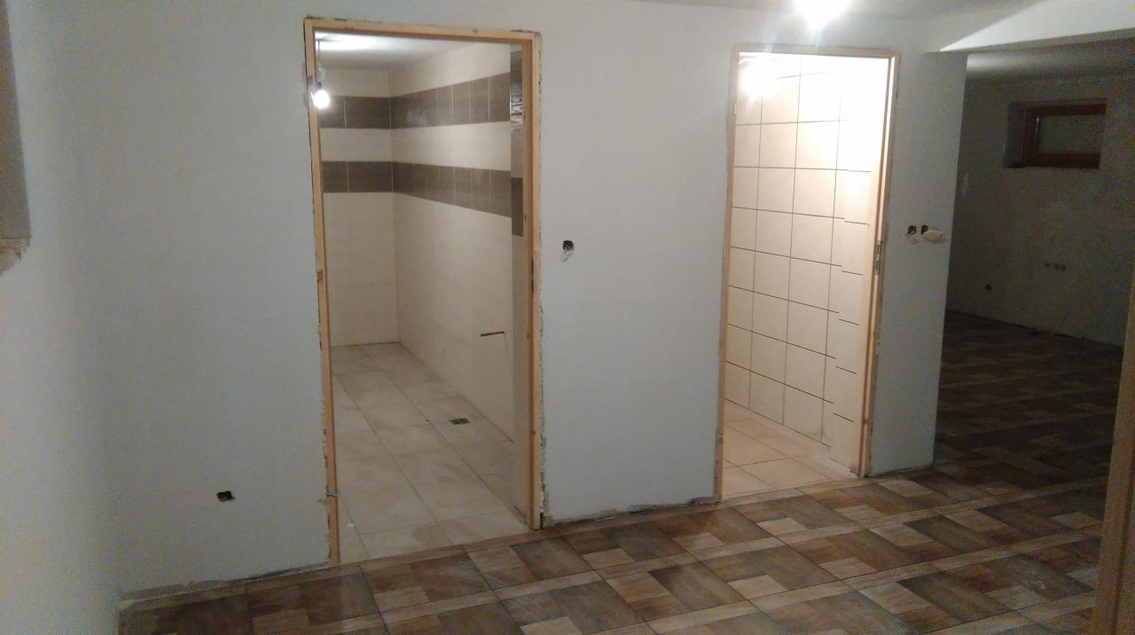 Dwa wejścia do łazienek z wykończonymi płytkami, widoczne surowe ościeżnice drzwiowe i fragment podłogi z płytek imitujących drewno. Widok na wnętrze w trakcie wykańczania.