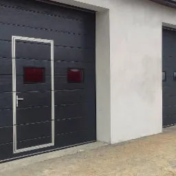 Szara segmentowa brama garażowa z drzwiami i małymi okienkami, widok na trzy bramy w rzędzie, jasny budynek.