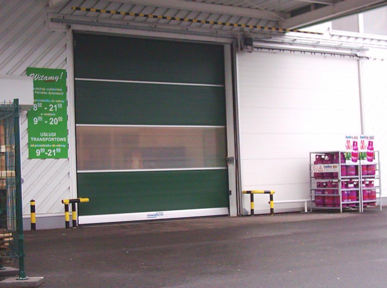 Zielona segmentowa brama garażowa Novoform z przeszklonym panelem, częściowo otwarta, z widocznym systemem prowadnic i stojakiem na butle gazowe obok.