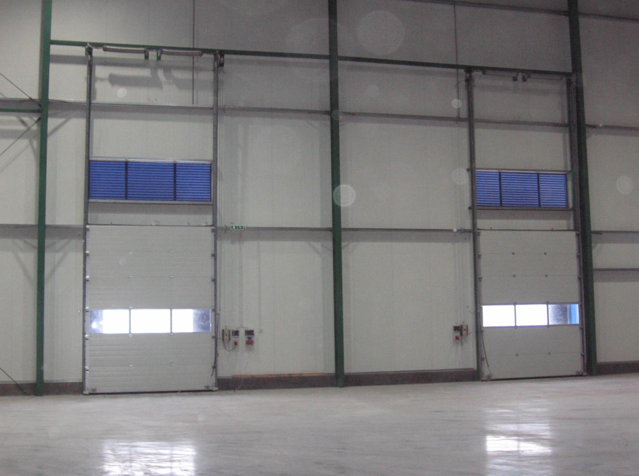 Dwie segmentowe bramy przemysłowe w hali, jedna częściowo otwarta, z widocznymi oknami i kratkami wentylacyjnymi, na tle białych ścian i betonowej podłogi.