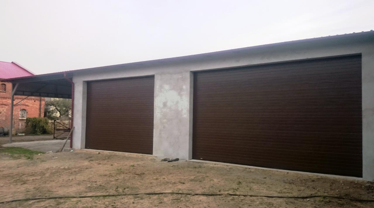 Dwie brązowe, segmentowe  do garażu w betonowym budynku. Po lewej wiata, w tle budynek z czerwonym dachem. Ziemiste podłoże.