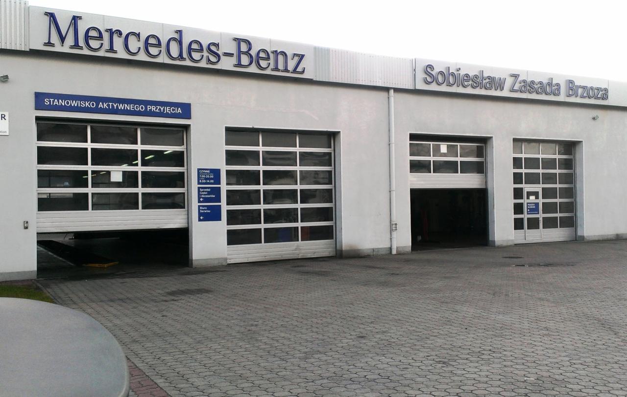 Biały budynek serwisu Mercedes-Benz z czterema bramami garażowymi, z których jedna jest częściowo otwarta, ukazując wnętrze warsztatu.