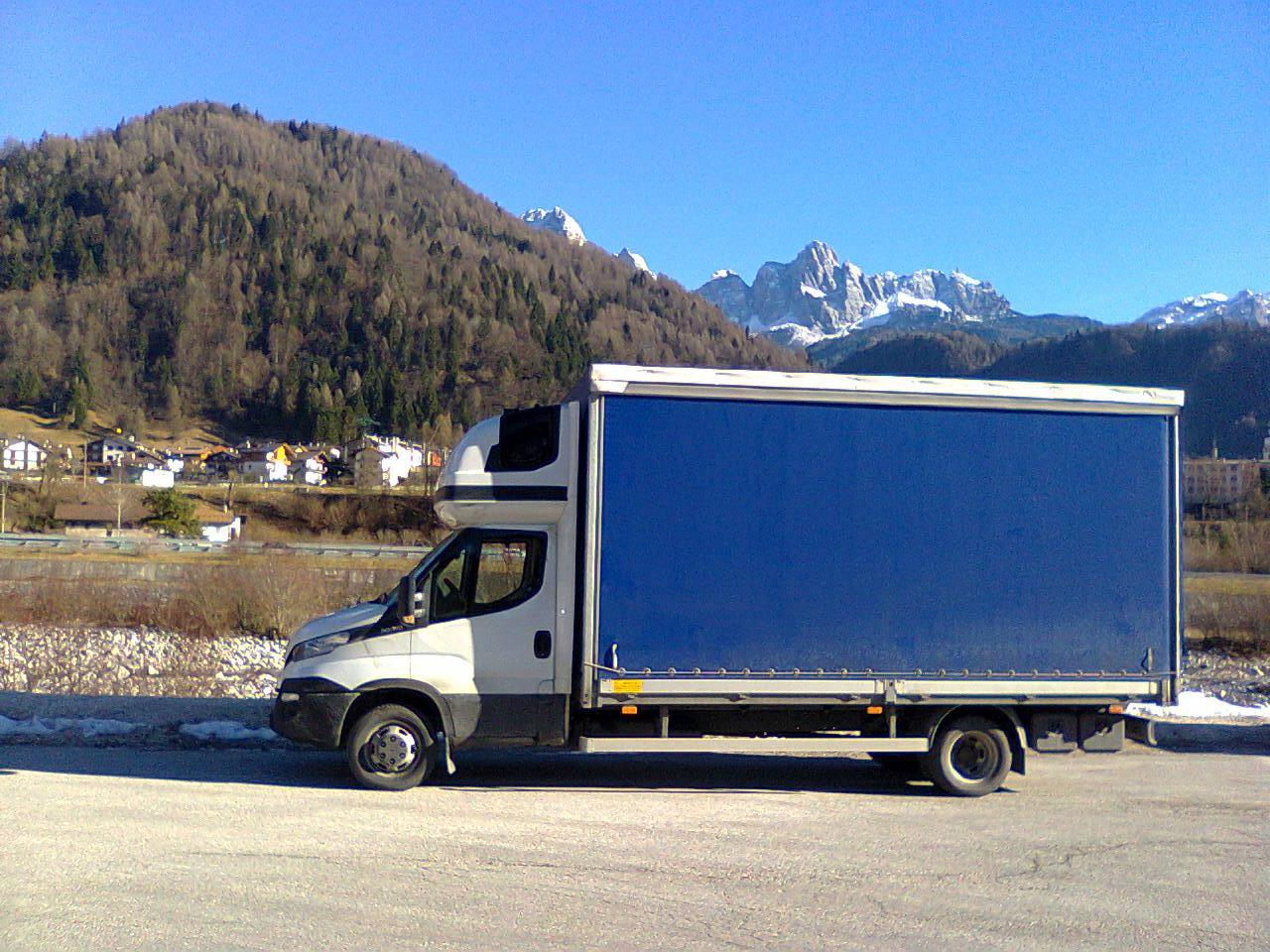 Ciężarówka z niebieską plandeką na tle górskiego krajobrazu z ośnieżonymi szczytami i zabudowaniami w dolinie.