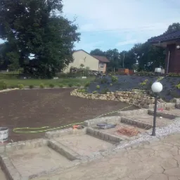 Prace ziemne i brukarskie w ogrodzie: wyrównany teren pod trawnik, kamienne schody z oświetleniem, narzędzia brukarskie na pierwszym planie.