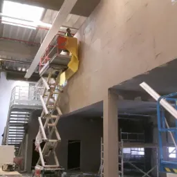 Prace wykończeniowe w hali: pracownik na podnośniku maluje ścianę. Widoczne rusztowania, materiały budowlane i surowy stan wnętrza. Hala w trakcie modernizacji.