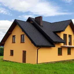 Żółty dom jednorodzinny z czarnym dachem na tle błękitnego nieba i zieleni. Widok z niskiej perspektywy, ukazujący detale elewacji i otoczenia.