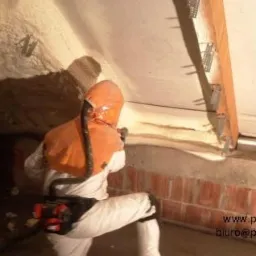 Pracownik w kombinezonie ochronnym aplikuje izolację natryskową na poddaszu, widoczne detale konstrukcyjne i fragment ocieplonej ściany z cegły.