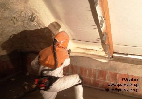 Pracownik w kombinezonie ochronnym aplikuje izolację natryskową na poddaszu, widoczne detale konstrukcyjne i fragment ocieplonej ściany z cegły.