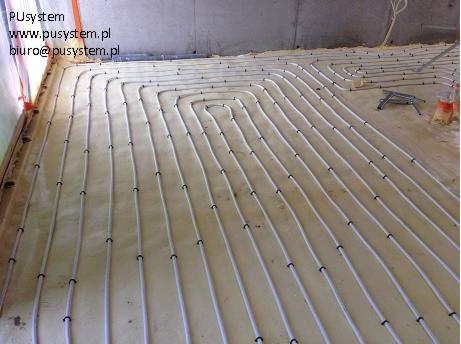 Instalacja ogrzewania podłogowego na izolacji z pianki. Białe rury ułożone w pętle, przymocowane czarnymi uchwytami. Widoczne narzędzia instalacyjne.