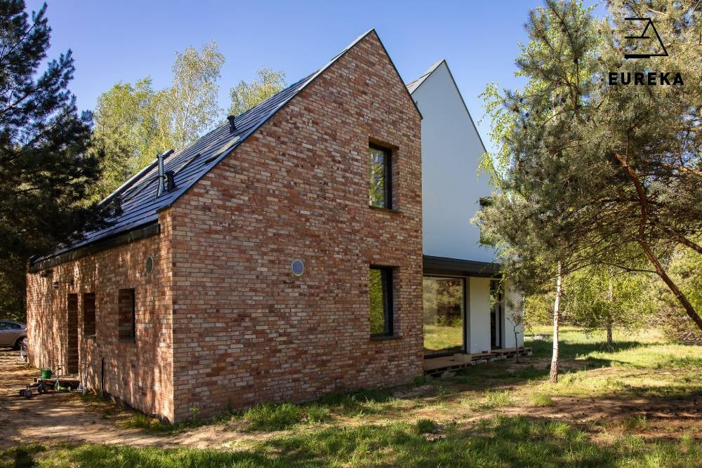 Nowoczesny dom jednorodzinny z cegły klinkierowej i białym tynkiem, z czarną dachówką i oknami, widoczny logo Eureka na tle drzew