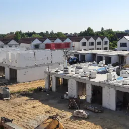 Budowa osiedla domów jednorodzinnych w stanie surowym otwartym, widok z góry na plac budowy z pracownikami i dostawczym samochodem na tle gotowych domów.