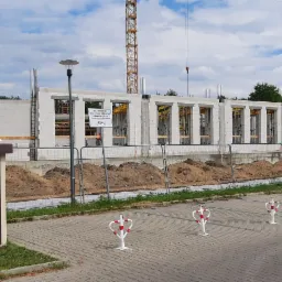 Wznoszenie ścian z betonu komórkowego na budowie domu jednorodzinnego, widoczne stalowe zbrojenia, szalunki okienne, oraz rozdzielnica elektryczna i zaparkowany samochód dostawczy w tle.