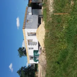 Wznoszenie dwukondygnacyjnego budynku z białych bloczków, widoczne drewniane szalunki okienne, hałda piasku i bujna trawa w otoczeniu, błękitne niebo z pojedynczymi obłokami.